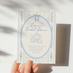May include: A light blue and cream bridal shower invitation with the text "Lauren's Bridal Shower". The invitation features a blue oval frame with a teapot and teacups illustration. The background has a striped pattern with bows and floral accents.