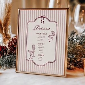 May include: A framed sign with a gold border features a drinks menu. The sign has a striped background with a decorative border and the word "Drinks" at the top. The menu lists cocktails, wine, and beers, with illustrations of a lemon and glasses.