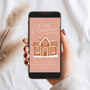 May include: A smartphone displays a digital invitation for a Christmas gathering. The invitation features a gingerbread house illustration with the text "A Cosy Christmas gathering". The background is a soft pink with falling snow.