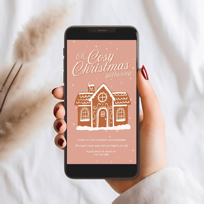 May include: A smartphone displays a digital invitation for a Christmas gathering. The invitation features a gingerbread house illustration with the text "A Cosy Christmas gathering". The background is a soft pink with falling snow.