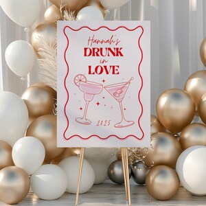 May include: A white sign with pink and red text that reads "Hannah's Drunk in Love 2025". The sign is decorated with two pink martini glasses and stars. The sign is on a gold easel.