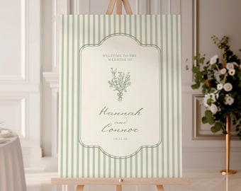 Puede incluir: Un letrero de bienvenida de boda sobre un caballete de madera. El letrero tiene un fondo a rayas verdes y blancas con un borde decorativo. El texto dice "Welcome to the Wedding of Hannah and Connor" con la fecha 10.11.26.
