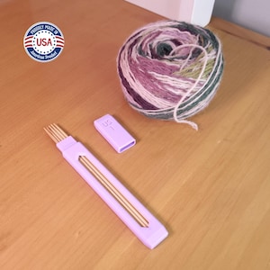 May include: A ball of multicolored yarn with shades of purple, green, and white sits next to a light purple knitting needle gauge and a case containing wooden knitting needles. The gauge is marked "US 1". The image also includes a "Proudly Made in USA" sticker.