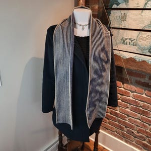 May include: A long, knitted scarf featuring a vertical striped pattern in dark blue and cream. The scarf is draped over a dark blue coat worn by a mannequin. A silver-toned necklace with a small pendant is visible around the neck of the mannequin.