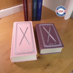 May include: Two knitting needle gauge books, one pink and one purple, with silver accents. Each book has an "X" design and reads "2mm - 8mm" and "US 0 - 11". The books are labeled "Notions" and "Proudly Made in USA".
