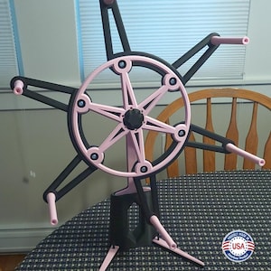 以下が含まれることがあります： 黒とピンクの糸巻き機。糸を玉に巻くための道具です。糸巻き機は、スポークのある中央の車輪が特徴で、脚付きのベースで支えられています。車輪とアームはピンク色で、残りの構造は黒色です。アメリカ製。