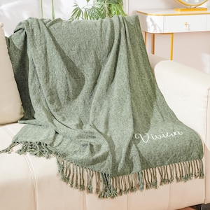 May include: A green throw blanket with fringe on a white sofa. The blanket has the name "Vivian" embroidered on it.