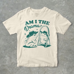 Peut inclure: Un t-shirt crème avec un graphique vert d'une grenouille portant un chapeau de cowboy et se regardant dans un miroir. Le texte "Am I the Drama?" est imprimé au-dessus de la grenouille.