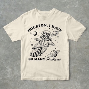Può includere: Una maglietta beige con una grafica nera di un procione che indossa una tuta da astronauta e galleggia nello spazio. Il testo "Houston, ho così tanti problemi" è stampato sulla maglietta.