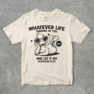 Pode incluir: Uma camiseta bege com um gráfico preto de um pato usando óculos de sol e o texto "Whatever life throws at you and let it hit someone else".