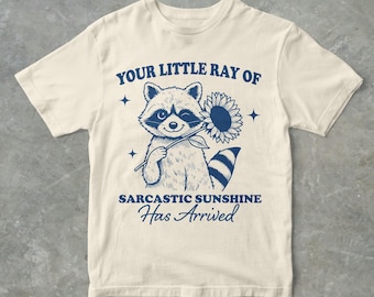 May include: A beige t-shirt featuring a blue graphic of a raccoon holding a sunflower. The text "Your Little Ray of Sarcastic Sunshine Has Arrived" is printed on the shirt.