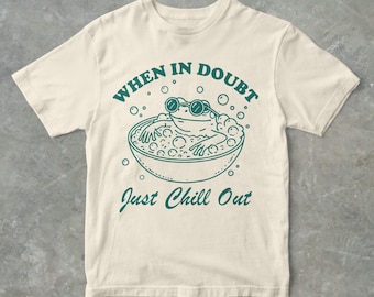 May include: A beige t-shirt featuring a green graphic of a frog wearing sunglasses and relaxing in a bubble bath. The text "When in doubt, just chill out" is printed below the frog.