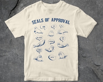 May include: A beige t-shirt featuring a graphic of seals with speech bubbles. The text on the shirt reads "Seals of Approval" and includes phrases like "AOK", "Yep", "Oh Yeah", "I Approve", "Copy That", "Roger Roger", and "Deffo".
