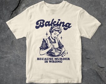 Puede incluir: Una camiseta beige con una ilustración retro de una mujer horneando. El texto "Baking because murder is wrong" está impreso en azul debajo de la ilustración.