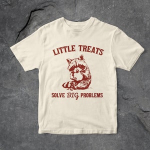 以下が含まれることがあります： ベージュのTシャツに、赤いアライグマのグラフィックと「Little Treats Solve Big Problems」というテキストがプリントされています。