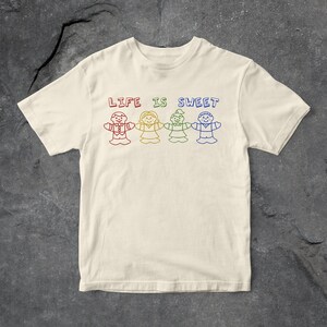 Op de afbeelding: Een wit T-shirt met de tekst "LIFE IS SWEET" bedrukt in regenboogkleuren. De tekst wordt omringd door vijf stickfiguren die elkaar vasthouden, ook in regenboogkleuren.