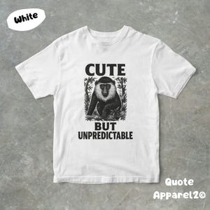 Op de afbeelding: Wit T-shirt met een zwarte afbeelding van een aap en de tekst "CUTE BUT UNPREDICTABLE." Het shirt heeft ook de tekst "Quote Apparel2O."
