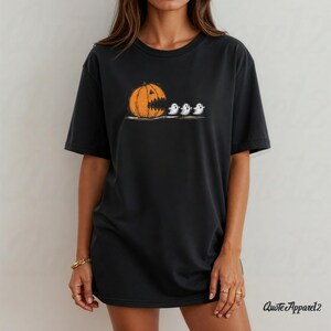 Op de afbeelding: Zwart T-shirt met een Halloween-thema. Het ontwerp toont een oranje pompoen met een open mond, gevolgd door drie kleine witte geesten. De tekst "Quote Apparel2" staat rechtsonder.