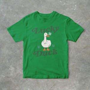 Op de afbeelding: Een groen T-shirt met een witte eend die een klavertje vier draagt. De tekst "Lucky Duck" staat op het shirt gedrukt.