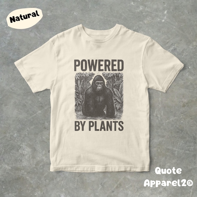 May include: Cream-colored t-shirt with a graphic of a gorilla in a jungle setting. The text "POWERED BY PLANTS" is printed above and below the gorilla image. The shirt has short sleeves and a crew neck.