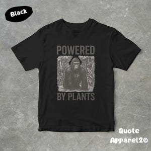 May include: Black t-shirt with a graphic of a gorilla in a jungle setting, and the text "POWERED BY PLANTS". The shirt has short sleeves and a crew neck. The brand name "Quote Apparel2o" is printed on the bottom right.