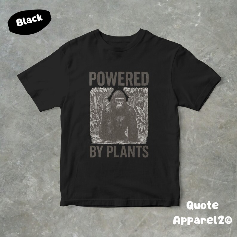 May include: Black t-shirt with a graphic of a gorilla in a jungle setting, and the text "POWERED BY PLANTS". The shirt has short sleeves and a crew neck. The brand name "Quote Apparel2o" is printed on the bottom right.