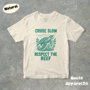 Op de afbeelding: Crèmekleurig T-shirt met een groene afbeelding van een zeeschildpad en de tekst "CRUISE SLOW RESPECT THE REEF." Het shirt heeft ook de tekst "Quote Apparel2o" en een "Natural" label.