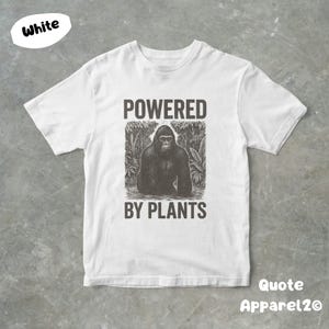 May include: White t-shirt with a black and white graphic of a gorilla in a jungle setting. The text "POWERED BY PLANTS" is above and below the gorilla image. The shirt is from Quote Apparel2O.