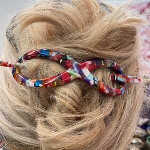May include: A colorful hair stick in an infinity shape, featuring a mix of red, blue, green, and white hues. The hair accessory is inserted into blonde hair, showcasing its decorative design. This hair accessory is a stylish addition to any hairstyle.
