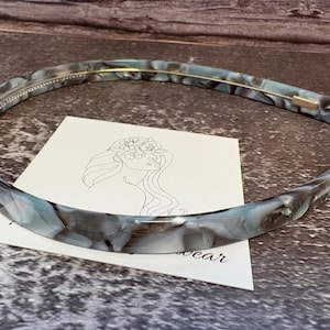May include: A thin, curved headband featuring a marbled pattern in shades of grey, white, and translucent blue. The headband has a smooth, polished finish and a thin gold-toned metal wire running along the inner edge for structural support.