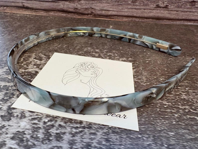 May include: A thin, curved headband featuring a marbled pattern in shades of grey, white, and translucent blue. The headband has a smooth, polished finish and a thin gold-toned metal wire running along the inner edge for structural support.