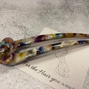 May include: A colorful hair clip with a floral design. The clip is made of plastic and has a white base with multi-colored swirls and flecks. The floral design is a large, multi-colored rose.
