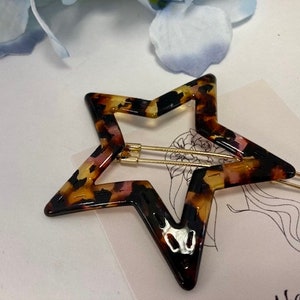 May include: A star-shaped hair clip with a tortoiseshell pattern in shades of brown, black, and pink. The clip has a gold-coloured metal clasp. The hair accessory is on a white surface with a floral design.