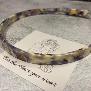 May include: A thin headband featuring a marbled pattern in shades of cream, purple, and brown. The headband rests on a white square card displaying a line drawing of a face with flowers and the printed text, For the Hair you wear.