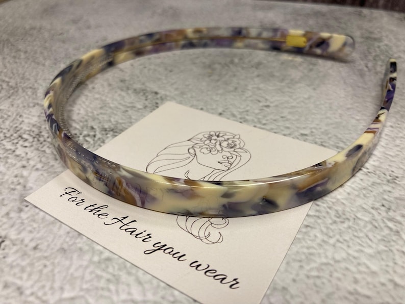May include: A thin headband featuring a marbled pattern in shades of cream, purple, and brown. The headband rests on a white square card displaying a line drawing of a face with flowers and the printed text, For the Hair you wear.