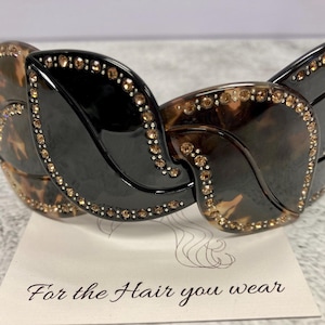 May include: A black and brown hair claw with rhinestones. The claw is shaped like a series of overlapping leaves. The text "For the Hair you wear" is printed on a white background.