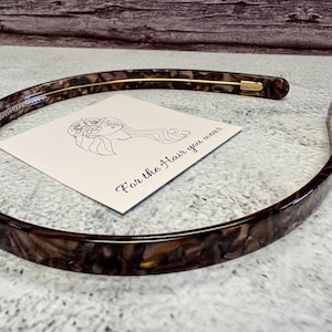 May include: A thin, curved headband featuring a brown and black tortoiseshell pattern with a smooth, glossy finish. A square white card is positioned inside the headband, displaying a black line drawing of a person and the text For the Hair you wear.