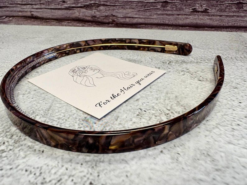 May include: A thin, curved headband featuring a brown and black tortoiseshell pattern with a smooth, glossy finish. A square white card is positioned inside the headband, displaying a black line drawing of a person and the text For the Hair you wear.