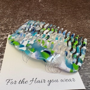 May include: A hair comb featuring a wavy, multi-coloured pattern of blue, green, and white acrylic. The comb rests on a white display card with a line drawing of a head and the printed text, For the Hair you wear, in a black script font.