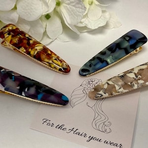 May include: Four colourful hair clips with gold metal clasps. The clips are made of patterned plastic and feature abstract designs in shades of brown, blue, purple, and white. The text "For the Hair you wear" is visible in the background.