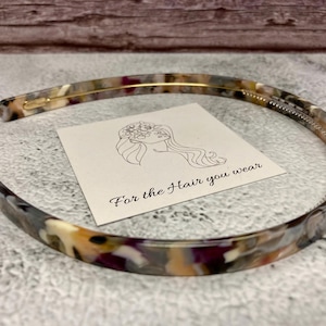 May include: A thin, semi-circular headband featuring a multi-colored tortoiseshell pattern in shades of brown, cream, and purple. A square white card is placed inside the headband, displaying a line drawing of a person and the text For the Hair you wear.