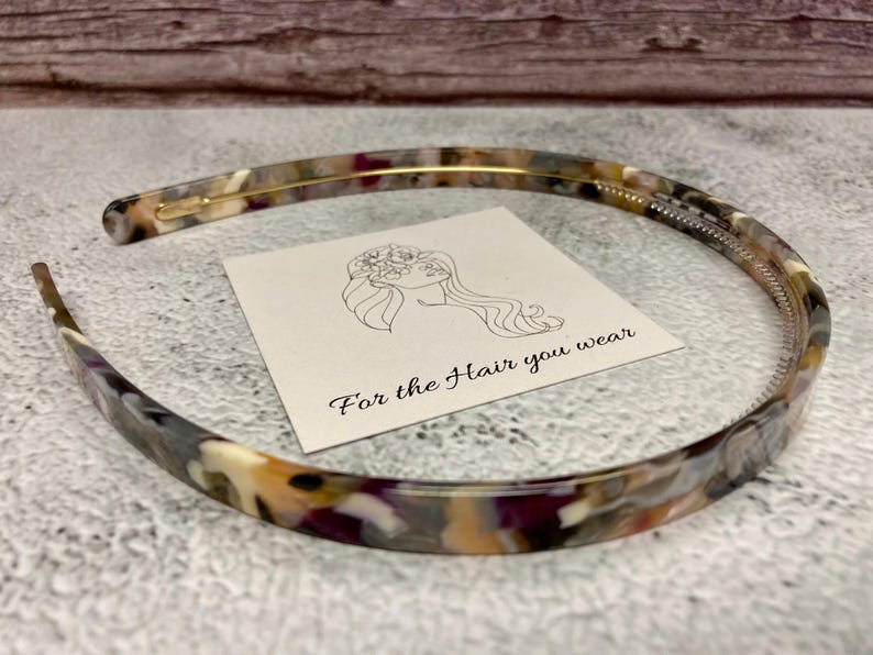 May include: A thin, semi-circular headband featuring a multi-colored tortoiseshell pattern in shades of brown, cream, and purple. A square white card is placed inside the headband, displaying a line drawing of a person and the text For the Hair you wear.