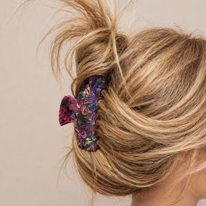Puede incluir: Una pinza para el pelo colorida con un diseño floral en tonos morado, rosa y verde. La pinza sujeta una parte del pelo rubio. El pelo está peinado en un moño.