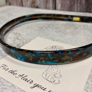 May include: A thin, flexible headband featuring a marbled pattern of deep blue, brown, and black tones. The headband has a smooth, glossy finish and rests on a white card with a line drawing of a hairstyle and the text For the Hair you w.