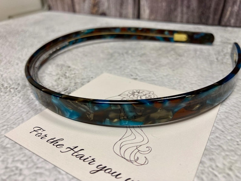 May include: A thin, flexible headband featuring a marbled pattern of deep blue, brown, and black tones. The headband has a smooth, glossy finish and rests on a white card with a line drawing of a hairstyle and the text For the Hair you w.