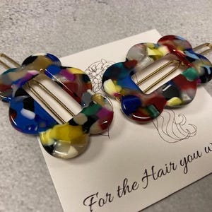 May include: Two colourful hair clips in the shape of a flower with a gold metal bar. The clips are made of a translucent plastic with a multi-coloured swirl pattern. The text "For the Hair you wear" is printed on a white card behind the clips.