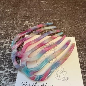 May include: A multi-coloured hair comb with a marbled pattern of pink, blue, and white hues. The comb features seven curved teeth and rests on a white display card featuring a line drawing of a person and the text For the Hair you wear.
