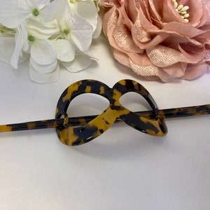 May include: A tortoiseshell hair stick with a decorative, infinity-shaped loop. The stick is made of brown and black plastic with a mottled pattern.
