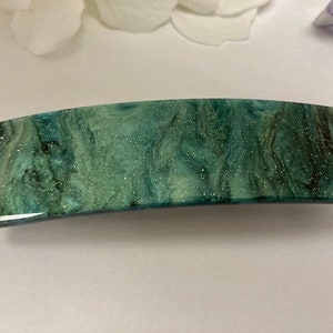 May include: A curved rectangular hair barrette in shades of teal, green, and dark brown, with a glittery, marbled pattern. The barrette is made from a smooth, shiny material.