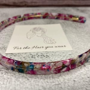 May include: A thin, circular headband featuring a multi-colored, marbled pattern with shades of magenta, white, blue, and brown. The headband is positioned around a square white card that displays a line drawing of a person and the text For the Hair you wear.
